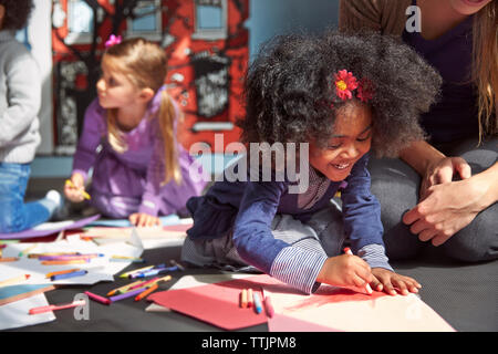 Cheerful girl drawing on paper while sitting by teacher at preschool Stock Photo