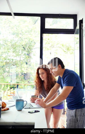 Couple watching laptop computer while having coffee at kitchen counter by window Stock Photo