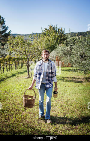 Portrait of man holding basket and wine bottle while standing on field in vineyard Stock Photo