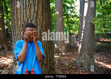 Playful boy covering eyes while standing against tree in forest Stock Photo