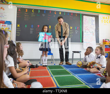 Girl explaining artwork while standing by teacher in classroom Stock Photo