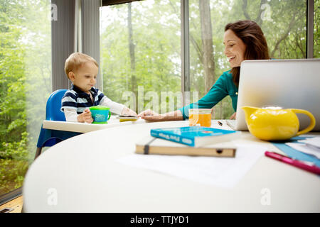 Smiling mother teaching son while using laptop at table against window Stock Photo