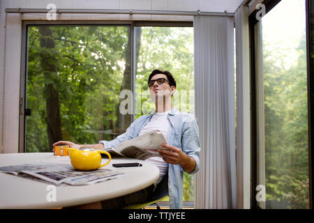 Thoughtful man holding newspaper while having tea at table against window Stock Photo