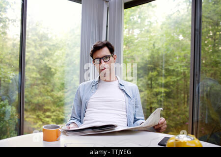 Portrait of man holding newspaper while having tea at table against window Stock Photo