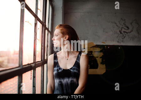 Thoughtful woman looking through glass window while standing against wall Stock Photo