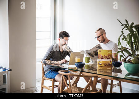 Homosexual couple using laptop and reading newspaper at breakfast table Stock Photo