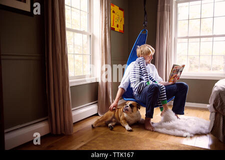 Father reading book to son and stroking dog while sitting on swing at home