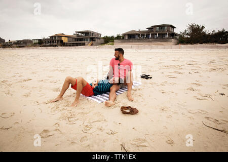 Happy homosexual couple resting on beach against sky Stock Photo