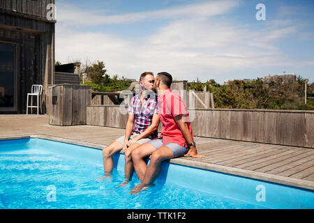 Homosexual couple kissing while sitting on poolside against sky Stock Photo