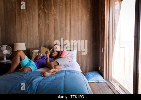 Gay men using tablet computer while lying on bed against wooden wall at resort Stock Photo