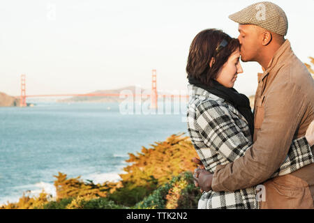 Man kissing girlfriend's forehead while embracing against bridge Stock Photo