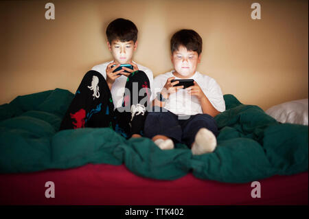 Brothers using phone while sitting on bed against wall at home Stock Photo
