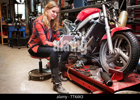 Woman using digital tablet while sitting on stool by bike at workshop Stock Photo