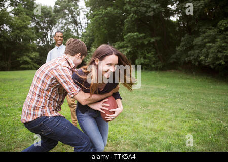 Man pulling back woman holding football ball while friend standing on grassy field Stock Photo