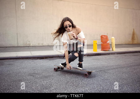 Young woman drinking juice while skateboarding on street Stock Photo