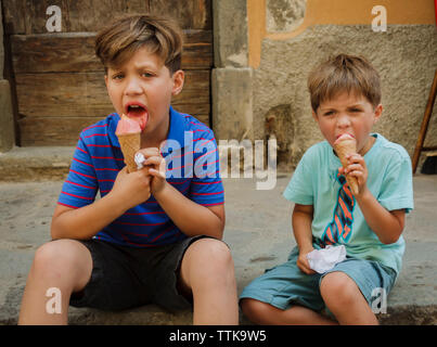 Brothers eating ice cream cones while sitting against wall Stock Photo