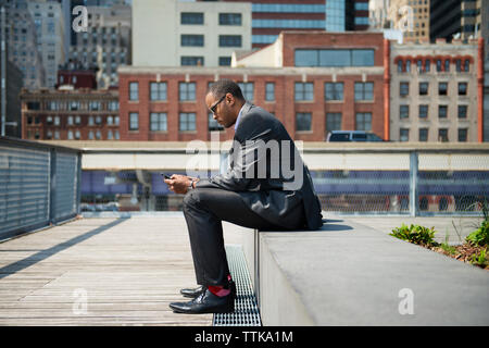 Side view of businessman using smart phone while sitting on retaining wall Stock Photo