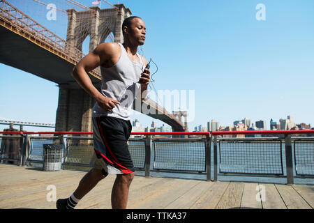 Man listening music and jogging on boardwalk against Brooklyn Bridge Stock Photo