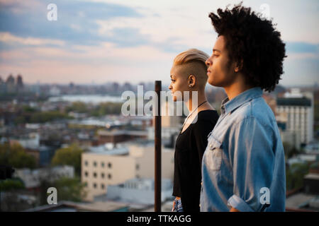 Thoughtful man and woman standing on rooftop against sky during sunset Stock Photo