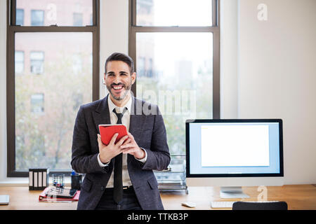 Happy businessman using tablet computer while leaning on desk in office Stock Photo