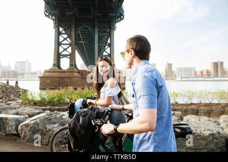 Happy couple walking with bicycle by Manhattan bridge against sky Stock Photo