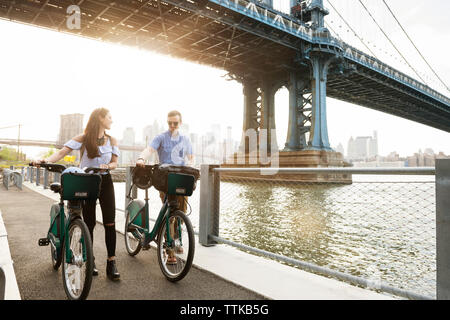 Couple walking with bicycles on promenade by East river against Manhattan bridge Stock Photo
