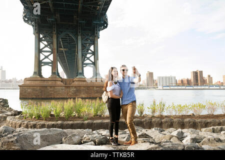 Couple taking selfie while standing on rocks under Manhattan Bridge against clear sky Stock Photo