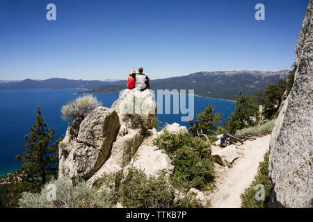 Couple sitting on cliff overlooking lake against clear blue sky Stock Photo