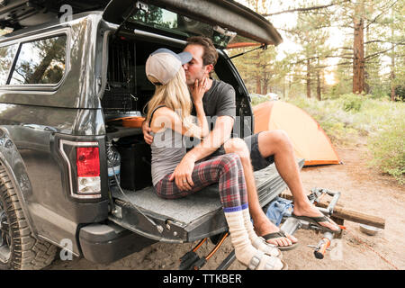 Romantic couple kissing while sitting on car trunk against trees in forest