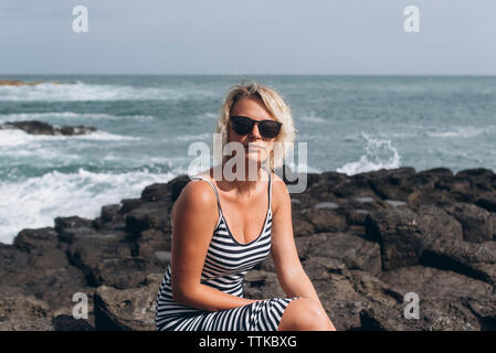 Portrait of confident woman wearing sunglasses while sitting on rocks at beach against sea Stock Photo