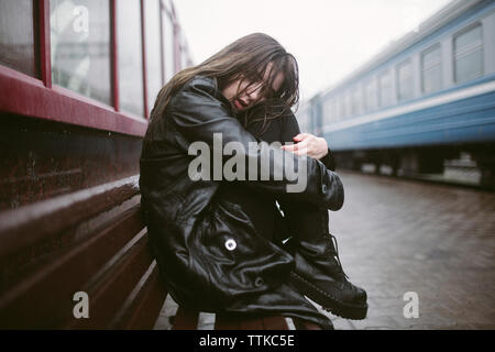 Portrait of sad woman sitting on bench at railroad station during rainy season Stock Photo