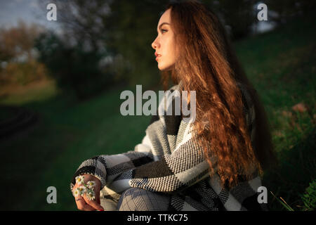 Thoughtful woman holding daisies while sitting on grassy field at park during sunset Stock Photo