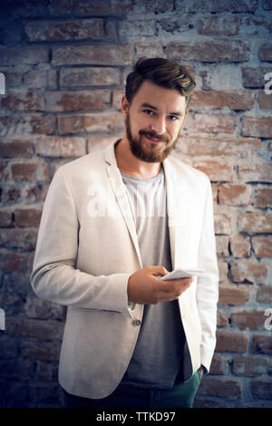 Portrait of businessman text messaging while standing against brick wall in office Stock Photo