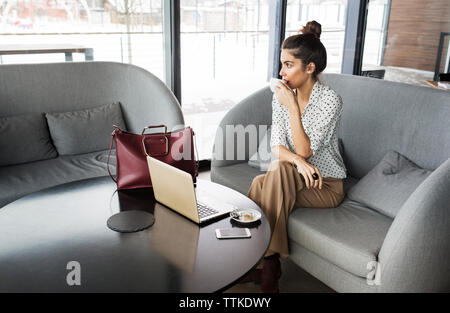 Thoughtful businesswoman drinking coffee while sitting on sofa at cafe Stock Photo