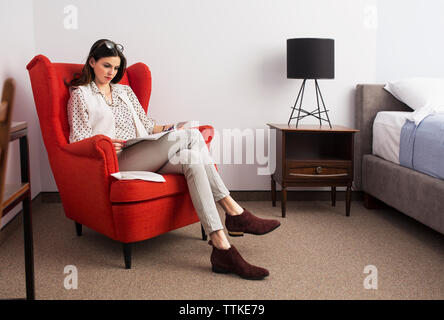 Businesswoman reading magazine while sitting on red armchair in hotel room Stock Photo