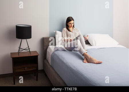 Businesswoman reading magazine while sitting on bed in hotel room Stock Photo