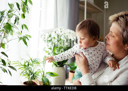 Mature woman carrying baby girl while standing by window at home Stock Photo