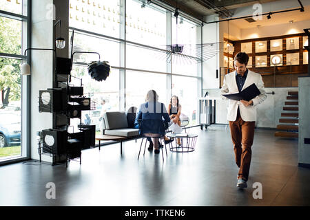 businessman reading file while walking in office by colleagues in background Stock Photo