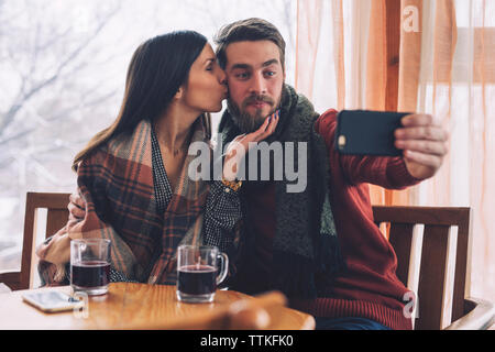 Woman kissing while boyfriend taking selfie at table in cafe Stock Photo