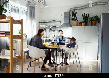 Parents looking at son sitting on high chair while having breakfast in kitchen Stock Photo