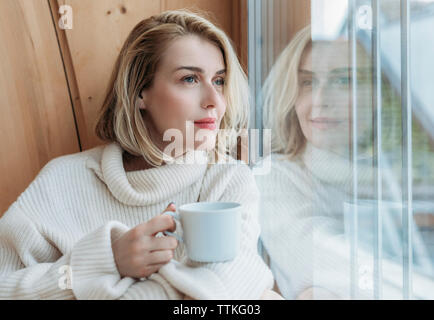 Woman holding coffee cup while looking through window at home Stock Photo