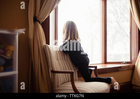 Rear view of girl kneeling on armchair by window at home Stock Photo