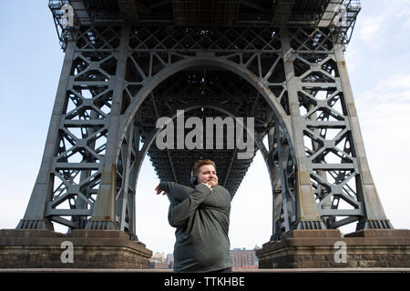 Overweight man stretching arms while standing below Williamsburg Bridge in city Stock Photo