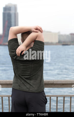 Rear view of overweight man stretching arms behind back while standing on bridge by river in city Stock Photo