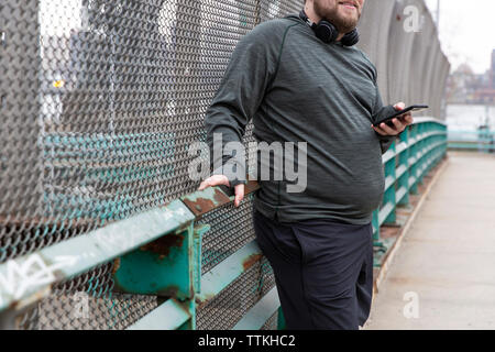 Midsection of overweight man holding smart phone while standing against metallic fence in city Stock Photo