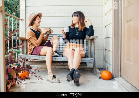 Cheerful female friends having drinks while sitting on wooden bench in porch Stock Photo
