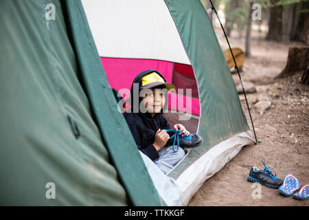 Portrait of boy holding shoes while sitting in tent at forest Stock Photo