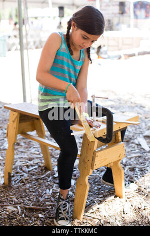 Girl shaping wood while sitting on bench at yard Stock Photo