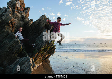 Playful boy jumping at beach while brother sitting on rocks