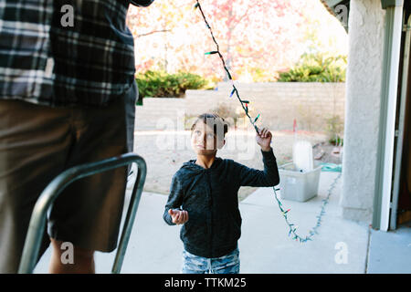 Son looking at father hanging colorful string lights on house during christmas Stock Photo